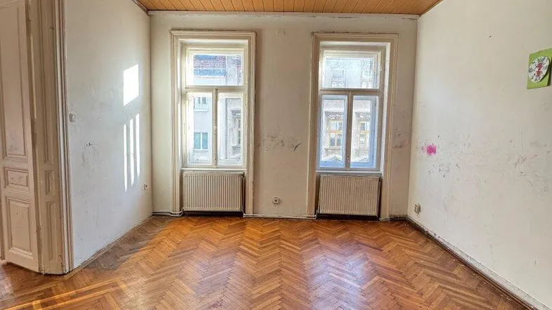 Helles Zimmer mit Fischgrätparkett, Holzdecke und zwei Fenstern, bereit zur Renovierung.