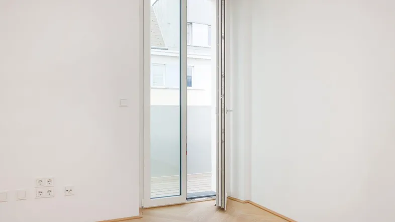 Helles Zimmer mit Fischgrätparkett und Zugang zu einem Balkon, ideal für Wohn- oder Schlafbereich.