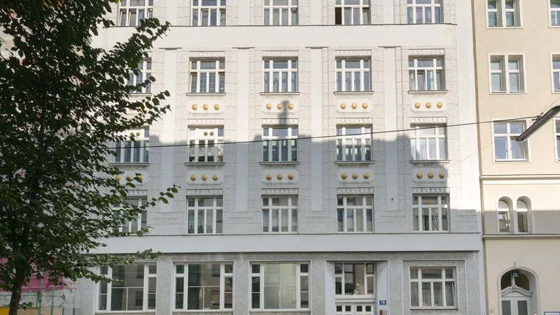Historische Fassade eines Mehrfamilienhauses mit vielen Fenstern und dekorativen Elementen an einer Stadtstraße.