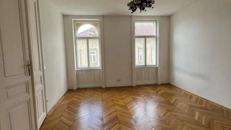Helles Zimmer mit klassischem Fischgrätparkett, hohen Decken und zwei großen Fenstern.