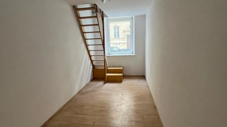 Schmaler Raum mit Holzleiter, die zu einer oberen Ebene führt, und einem Fenster am Ende.