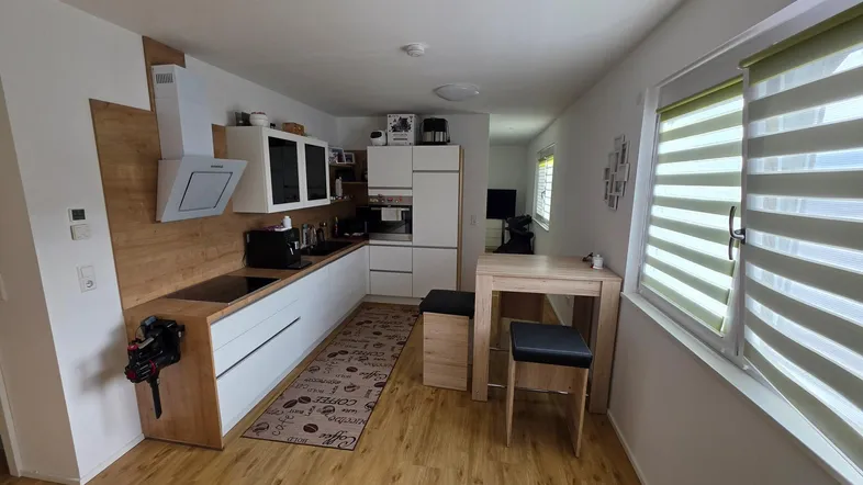 Moderne Küche mit weißen Fronten, Holzdekor und einem kleinen Esstisch am Fenster.