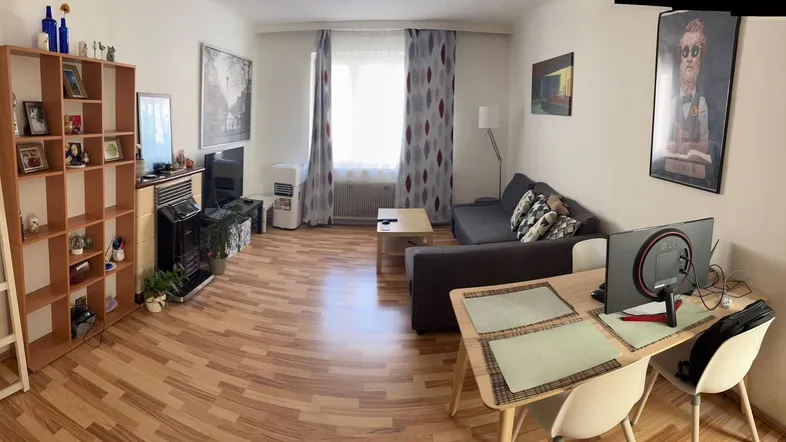 Geräumiges Wohnzimmer mit Holzboden, Sofa, Schreibtisch und Bücherregal, hell durch großes Fenster.