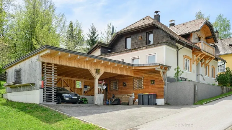 Traditionelles Haus mit großem Holzcarport und Stellplätzen, umgeben von grüner Landschaft.