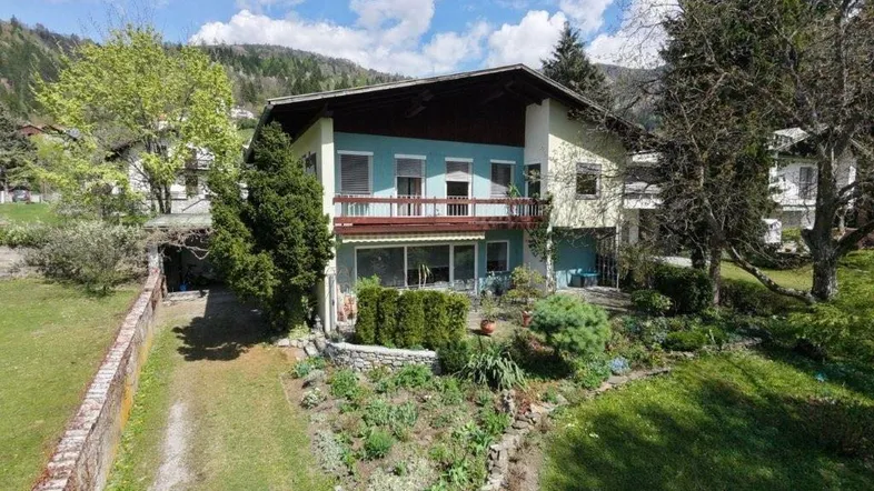 Großzügiges Einfamilienhaus mit blau-grüner Fassade, Balkon und gepflegtem Vorgarten in bergiger Umgebung.