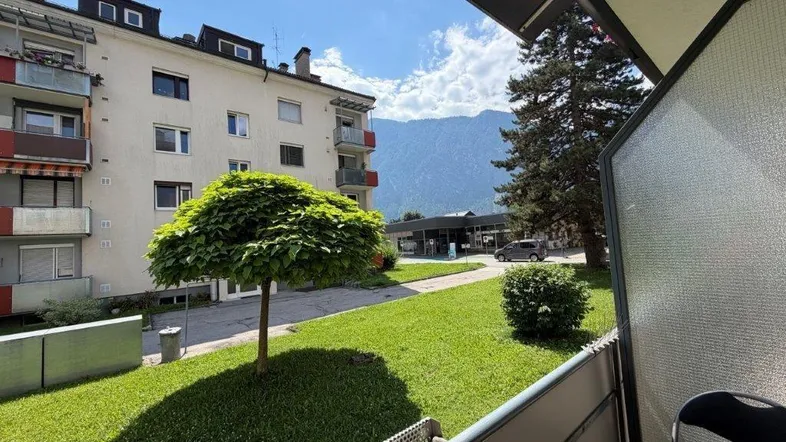Blick vom Balkon auf die grüne Umgebung, ein Mehrfamilienhaus und die Berge im Hintergrund.