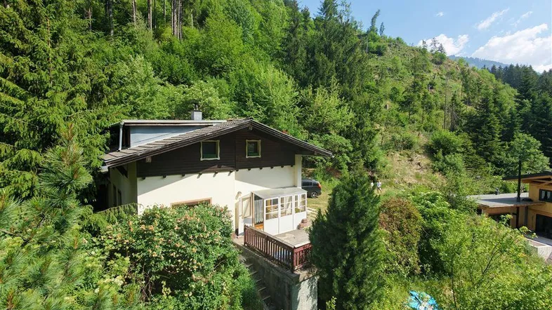 Haus in Hanglage umgeben von dichtem Wald und Bergen, mit kleiner Terrasse und Wintergarten.