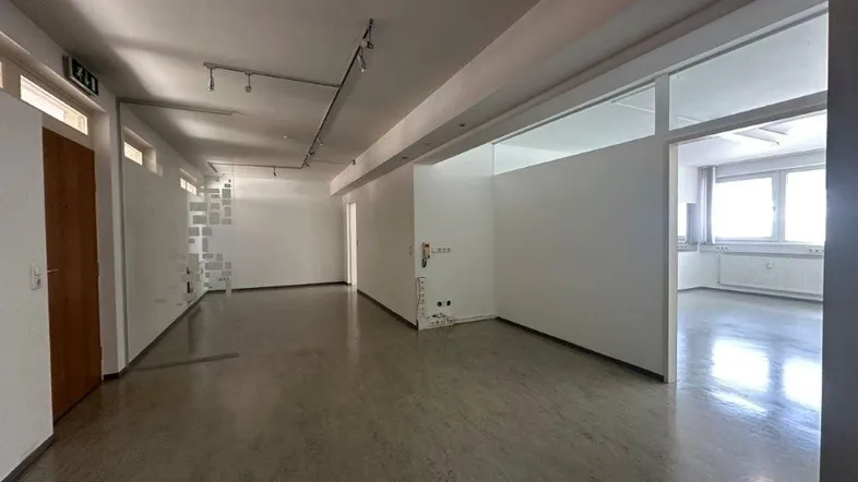 Geräumiger Büroraum mit hellen Wänden, Linoleumboden und Blick in einen weiteren Bereich.