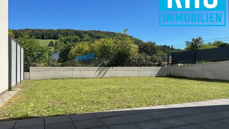 Gepflegter Gartenbereich mit Rasenfläche und gepflasterter Terrasse, umgeben von einer Betonmauer.