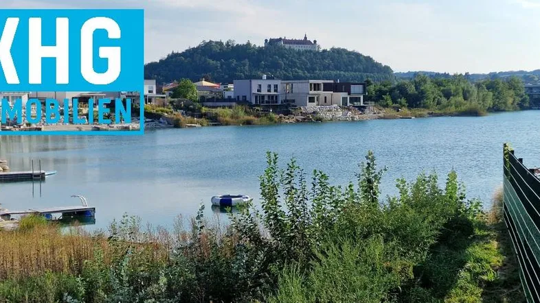 Seeblick mit modernen Häusern am Ufer und einer Burg auf dem Hügel im Hintergrund.