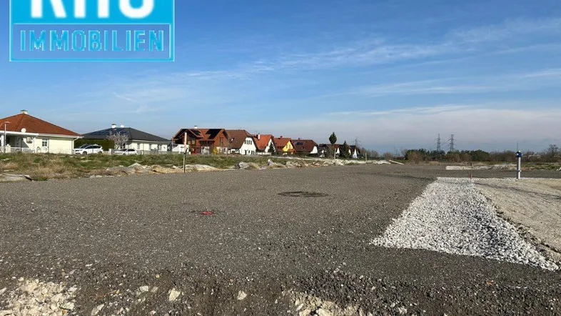 Großes Baugrundstück mit Schotterfläche und Häusern im Hintergrund unter blauem Himmel.