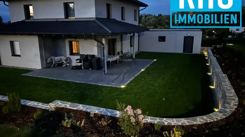 Modernes Einfamilienhaus bei Dämmerung mit beleuchtetem Garten und Terrasse.