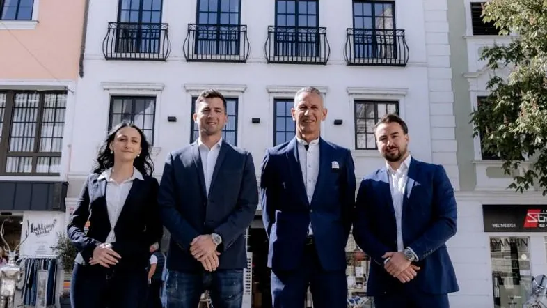 Moderne weiße Fassade eines Gebäudes mit schwarzen Balkonen und vier Personen davor.