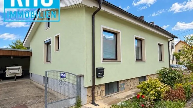Grünes Einfamilienhaus mit Vorgarten und Einfahrt unter blauem Himmel.