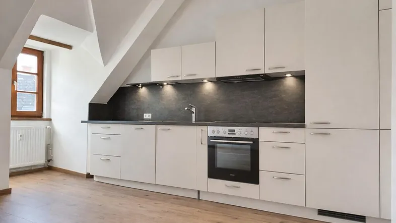 Helle Einbauküche im Dachgeschoss mit weißen Schränken, dunkler Arbeitsplatte und integriertem Backofen.