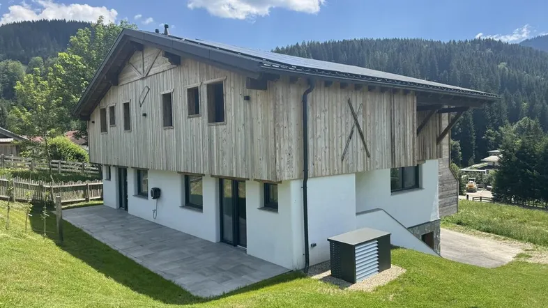Modernes Chalet mit Holzfassade, großer Terrasse und Photovoltaikanlage in alpiner Umgebung.