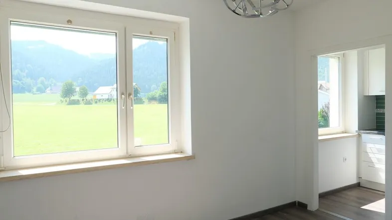 Heller Wohnbereich mit großem Fenster, Blick ins Grüne und Zugang zur Küche.