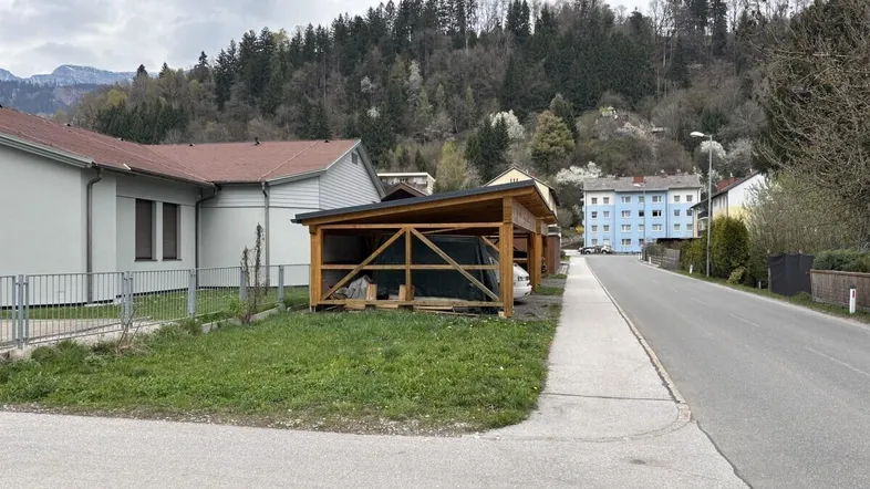 Blick auf eine Wohnstraße mit einem Holzcarport und Häusern im Hintergrund, umgeben von Hügeln.