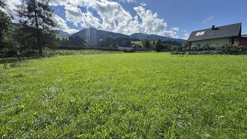 Weitläufige Grünfläche mit Bergblick und einem modernen Haus im Hintergrund unter blauem Himmel.