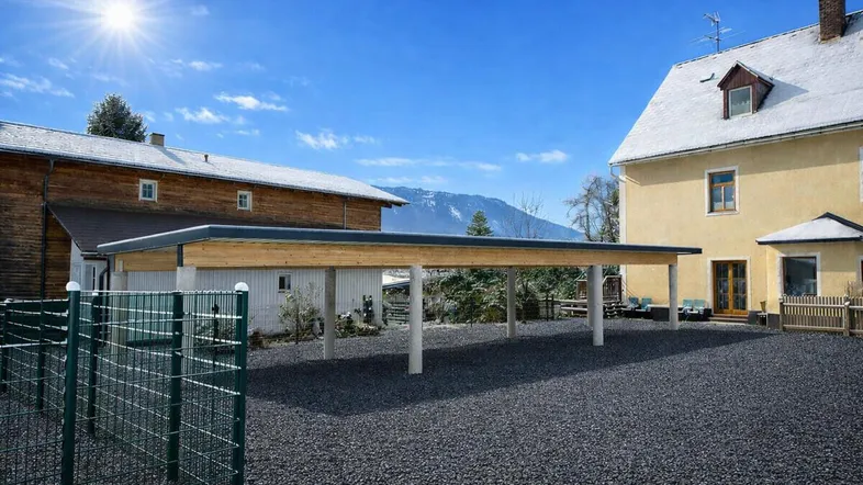 Moderner Carport mit Holzbalken und Betonsäulen vor einem Gebäude mit Bergblick an einem sonnigen Tag.