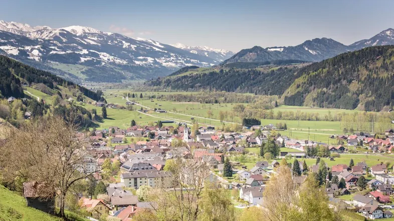 Panoramablick auf ein malerisches Dorf in einem grünen Tal mit schneebedeckten Bergen im Hintergrund.