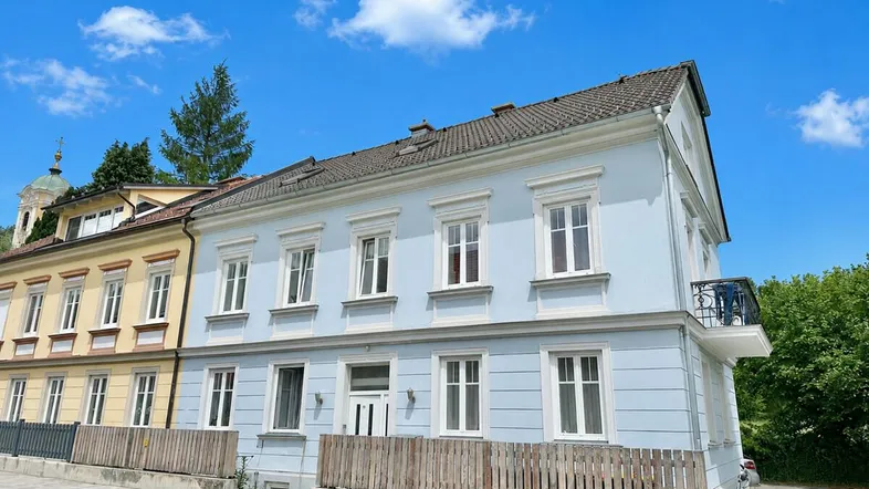 Helles Gebäude mit hellblauer Fassade, weißen Fenstern und kleinem Balkon unter blauem Himmel.
