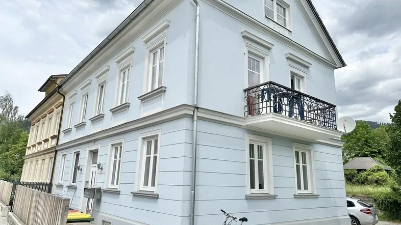 Helles Mehrfamilienhaus mit klassischer Architektur, Balkon und Satteldach unter bewölktem Himmel.