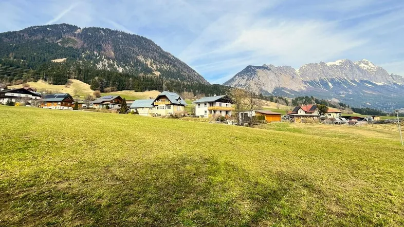 Weitläufige grüne Landschaft mit traditionellen Häusern und majestätischem Bergblick im Hintergrund.