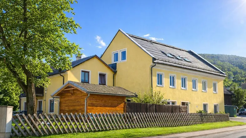 Gelbes Mehrfamilienhaus mit Holzzaun und grüner Umgebung unter blauem Himmel.