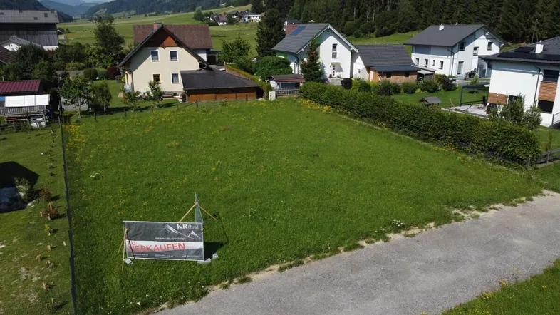 Luftaufnahme eines grünen Baugrundstücks mit umliegenden Häusern und Bergblick.