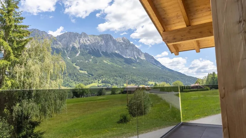 Blick von einem modernen Balkon mit Glasbrüstung auf die umliegende Berglandschaft und grüne Wiesen.