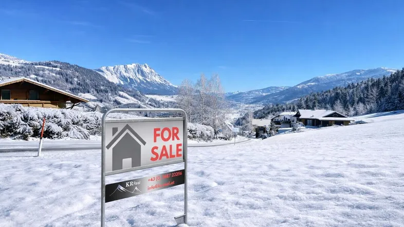 Verkaufsschild auf einem schneebedeckten Grundstück mit Alpenpanorama im Hintergrund.