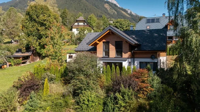 Modernes Haus mit Holzfassade und Satteldach, eingebettet in eine grüne Landschaft mit Bergblick.