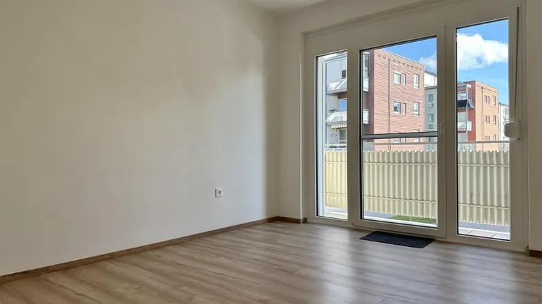 Helles, leeres Zimmer mit Laminatboden und Zugang zu einem kleinen Balkon.