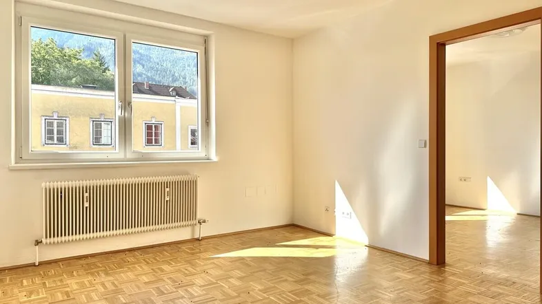 Helles, leeres Zimmer mit Parkettboden und großem Fenster, das einen Blick auf die Berge und umliegende Gebäude bietet.
