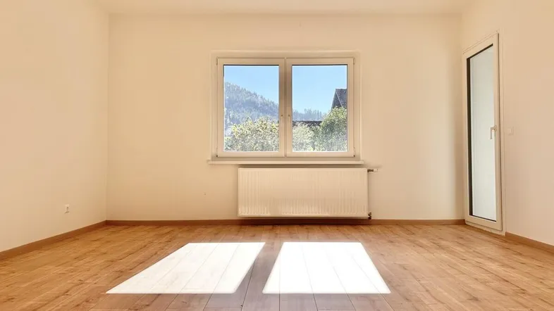 Helles, leeres Zimmer mit Holzboden, großem Fenster und Zugang zu einem Balkon.