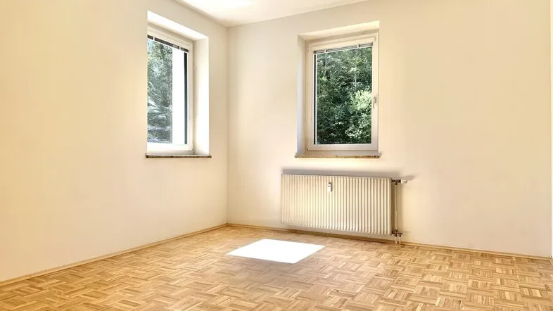 Helles Zimmer mit Parkettboden und zwei Fenstern, die viel Tageslicht hereinlassen.