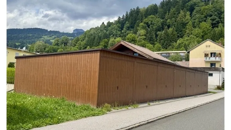 Lange, braune Holzfassade eines Gebäudes mit bewaldeten Hügeln im Hintergrund.