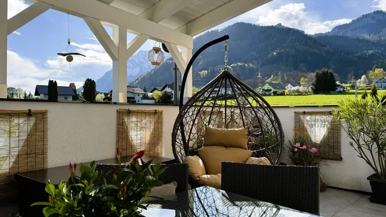 Gemütliche Terrasse mit Hängesessel und Esstisch, umgeben von grünen Bergen.