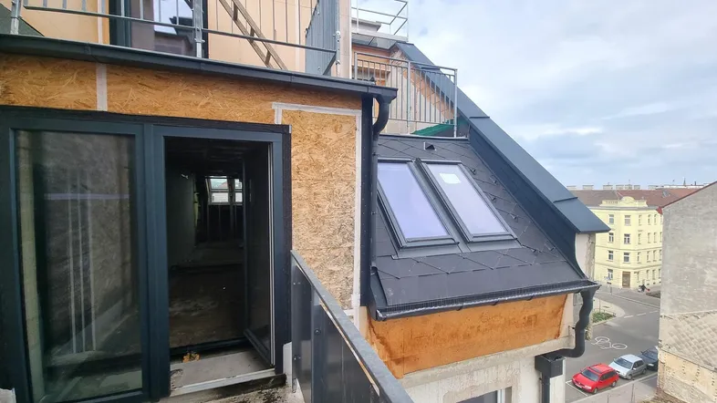Kleiner Balkon im Bau mit Metallgeländer und Blick auf die Stadt, angrenzend an ein Dachfenster.
