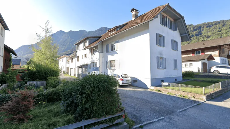 Weißes Wohngebäude mit Satteldach und hellen Fensterläden, umgeben von grüner Landschaft und Bergen.