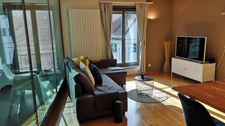 Helles Wohnzimmer mit bequemer Couch, Fernseher und Zugang zu einem Balkon oder einer Loggia.