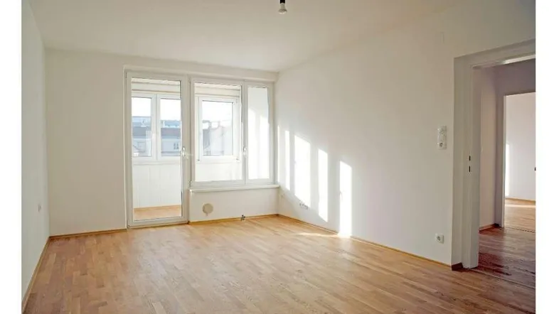 Helles, leeres Zimmer mit Parkettboden und Zugang zu einem Balkon, durchflutet von natürlichem Licht.