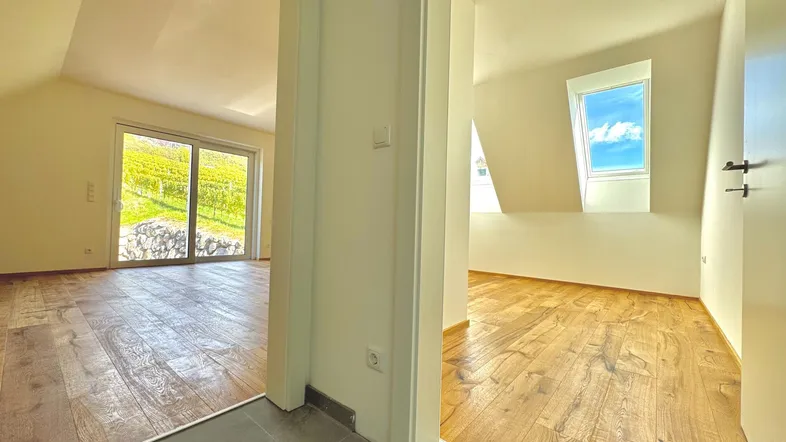 Heller Dachgeschossbereich mit Holzboden, Schrägen und Blick auf die Weinberge durch große Fenster.