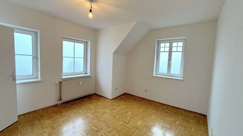 Helles Schlafzimmer im Dachgeschoss mit Parkettboden und mehreren Fenstern, die viel Licht hereinlassen.