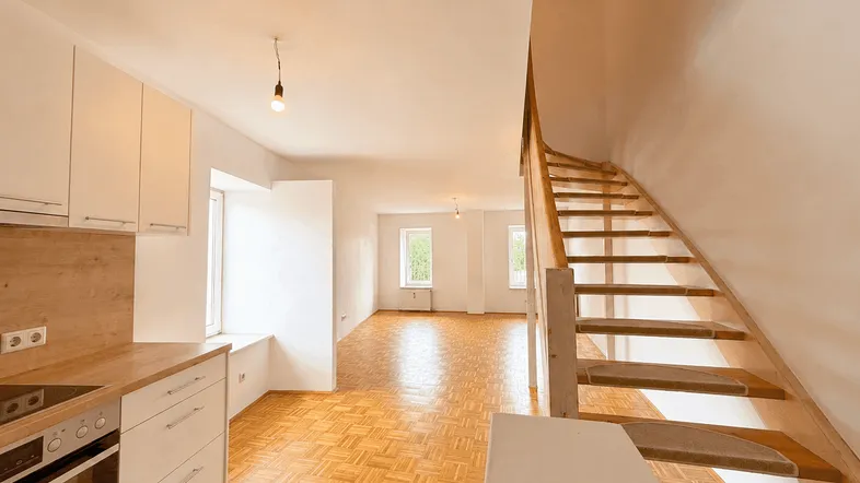 Heller offener Wohnbereich mit integrierter Küchenzeile, Parkettboden und Holztreppe zum Obergeschoss.