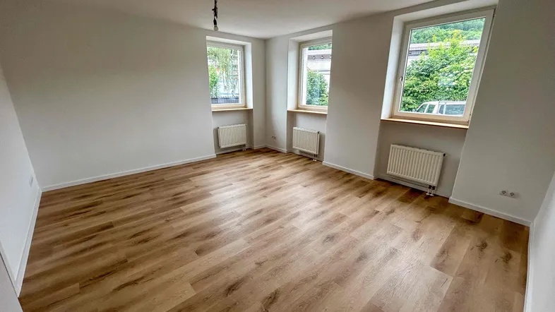 Helles, leeres Wohnzimmer mit neuem Laminatboden und drei Fenstern, die viel Tageslicht hereinlassen.