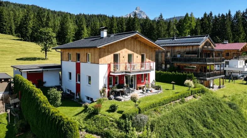 Modernes Haus mit Holzfassade und weißem Putz, umgeben von grünen Wiesen und Bergen.