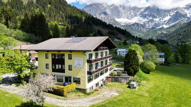 Luftaufnahme eines gelben Gebäudes mit Balkonen und Solarpaneelen, umgeben von grünen Wiesen und Bergen.