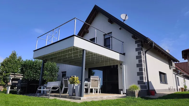 Modernes Haus mit großer überdachter Terrasse, Balkon und gepflegtem Rasen unter blauem Himmel.
